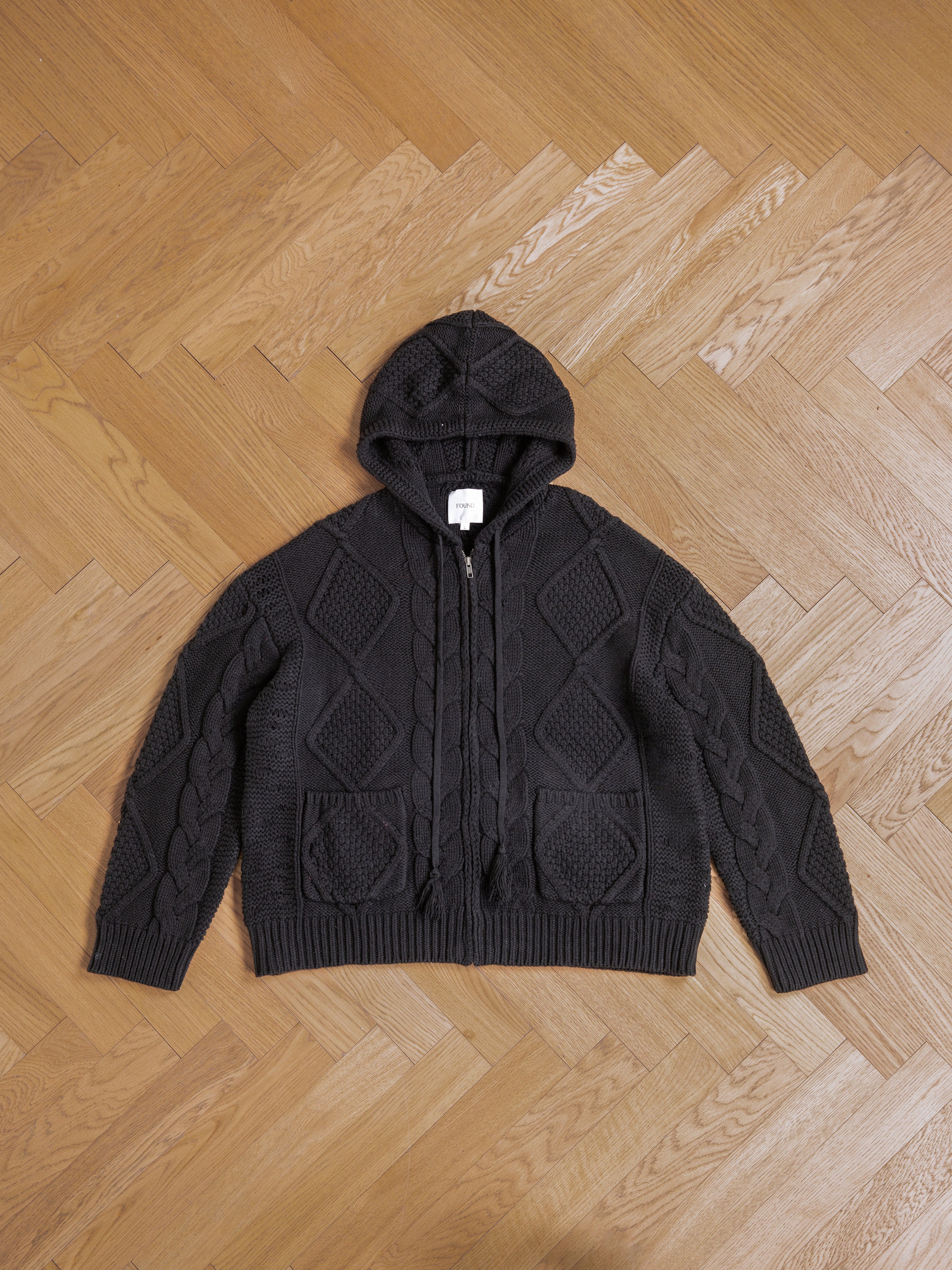 Black chunky knit Found Zip Up Cable Knit Hoodie with a diamond cable knit pattern, placed flat on a parquet wooden floor, offering both comfort and warmth.