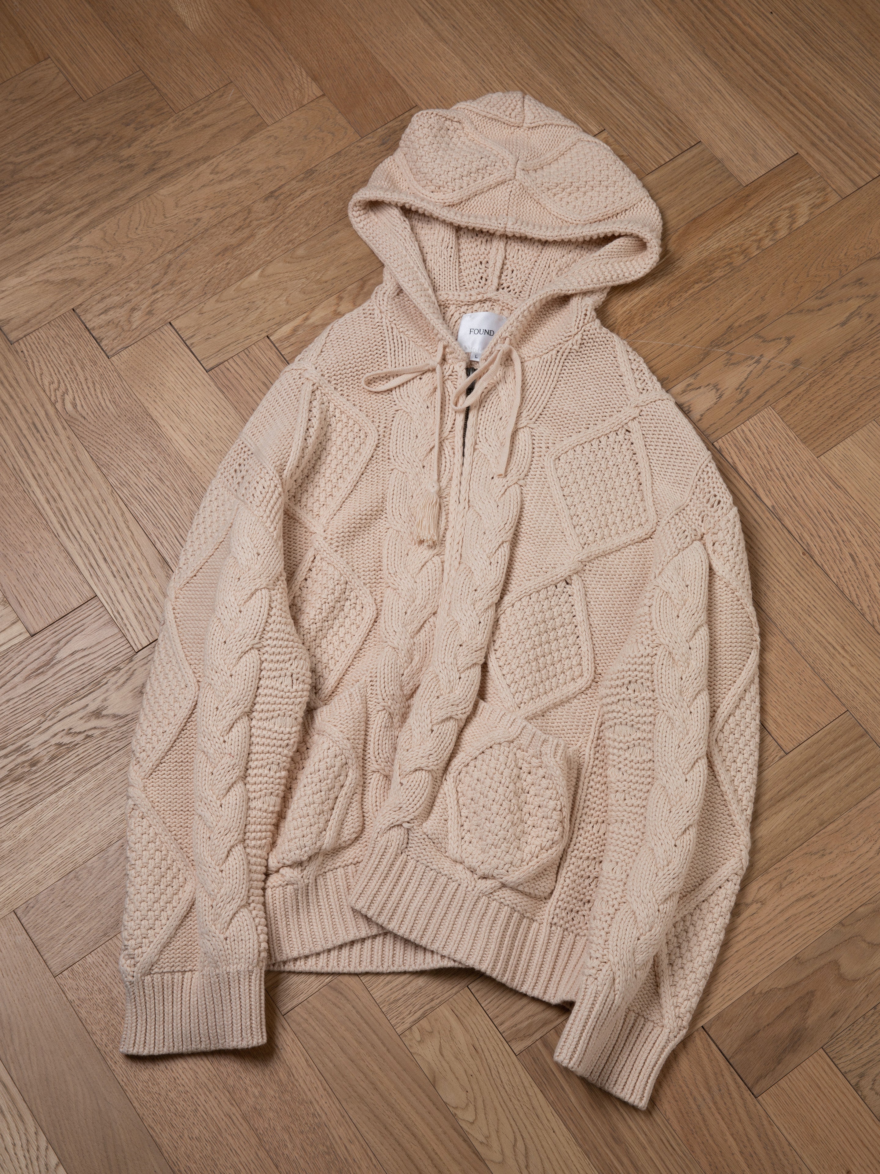 A beige, chunky knit hoodie with a front pocket and drawstrings is laid flat on a wooden floor, exuding comfort and warmth. The "Zip Up Cable Knit Hoodie" by Found showcases cozy sophistication in its design.