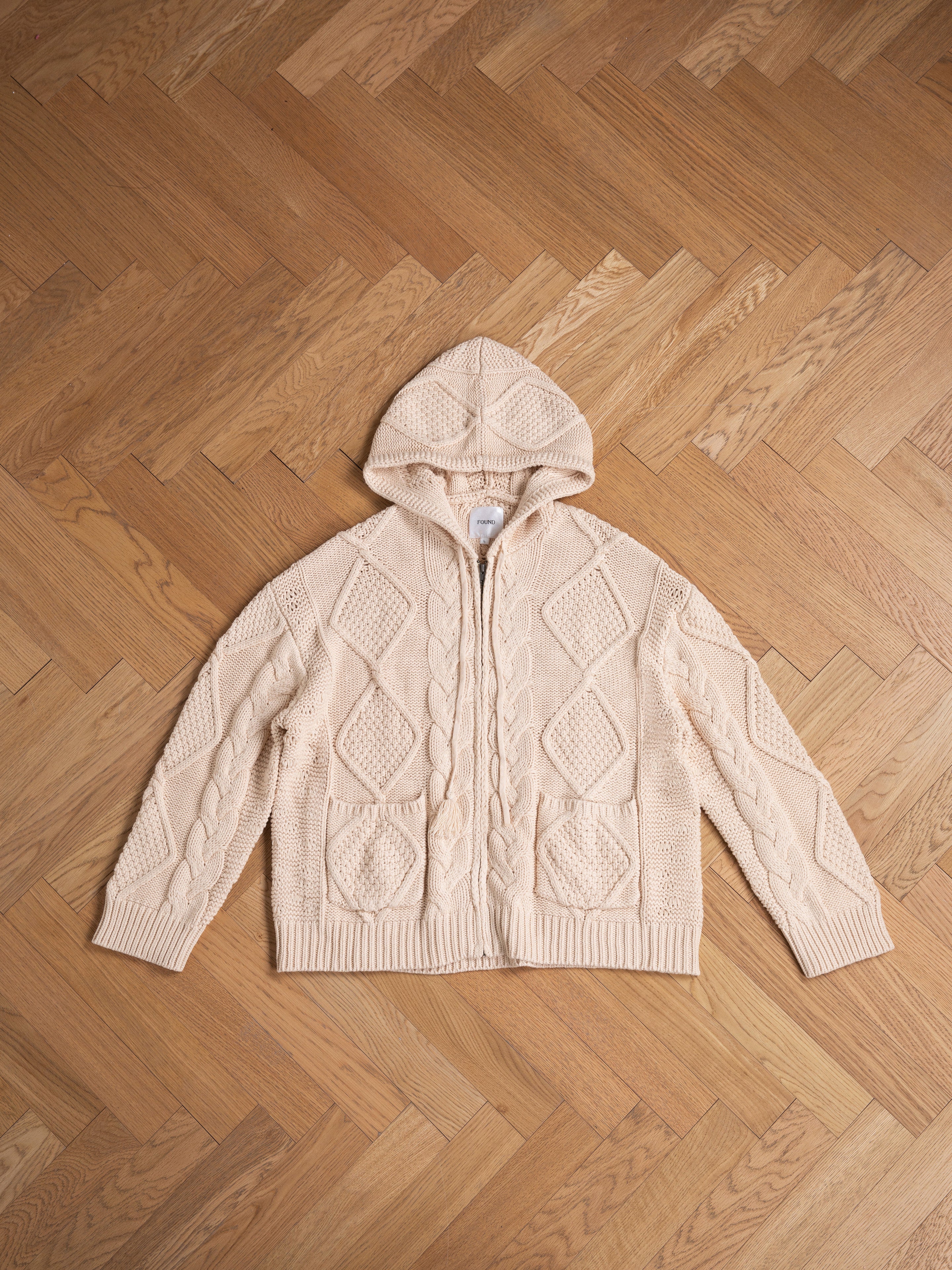 A cream chunky-knit Zip Up Cable Knit Hoodie by Found with a diamond and cable pattern design, laid flat on a wood herringbone floor, promises comfort and warmth.