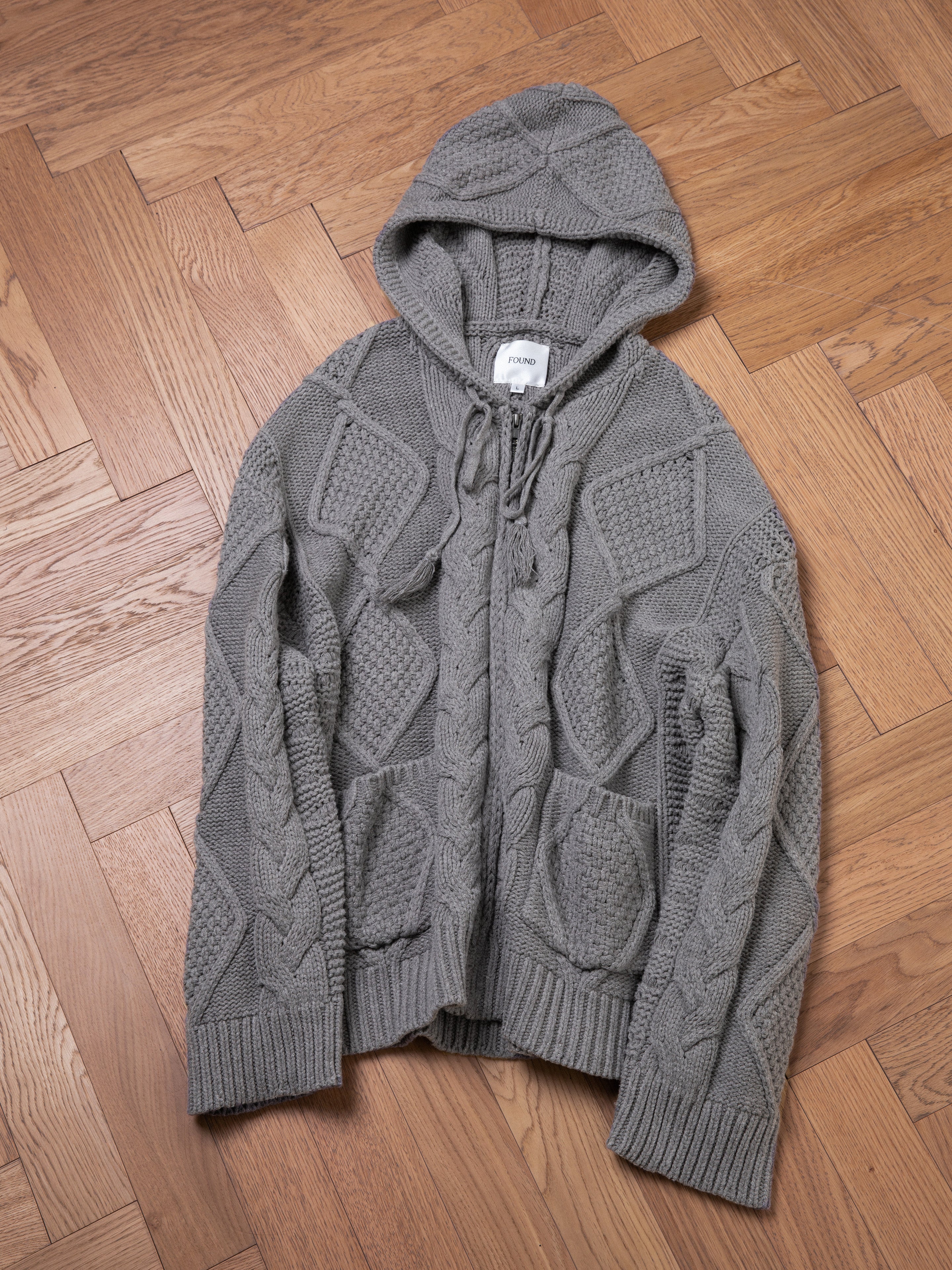 A gray Found Zip Up Cable Knit Hoodie with a chunky knit design, featuring a drawstring hood and front pockets, is laid flat on a wooden parquet floor.