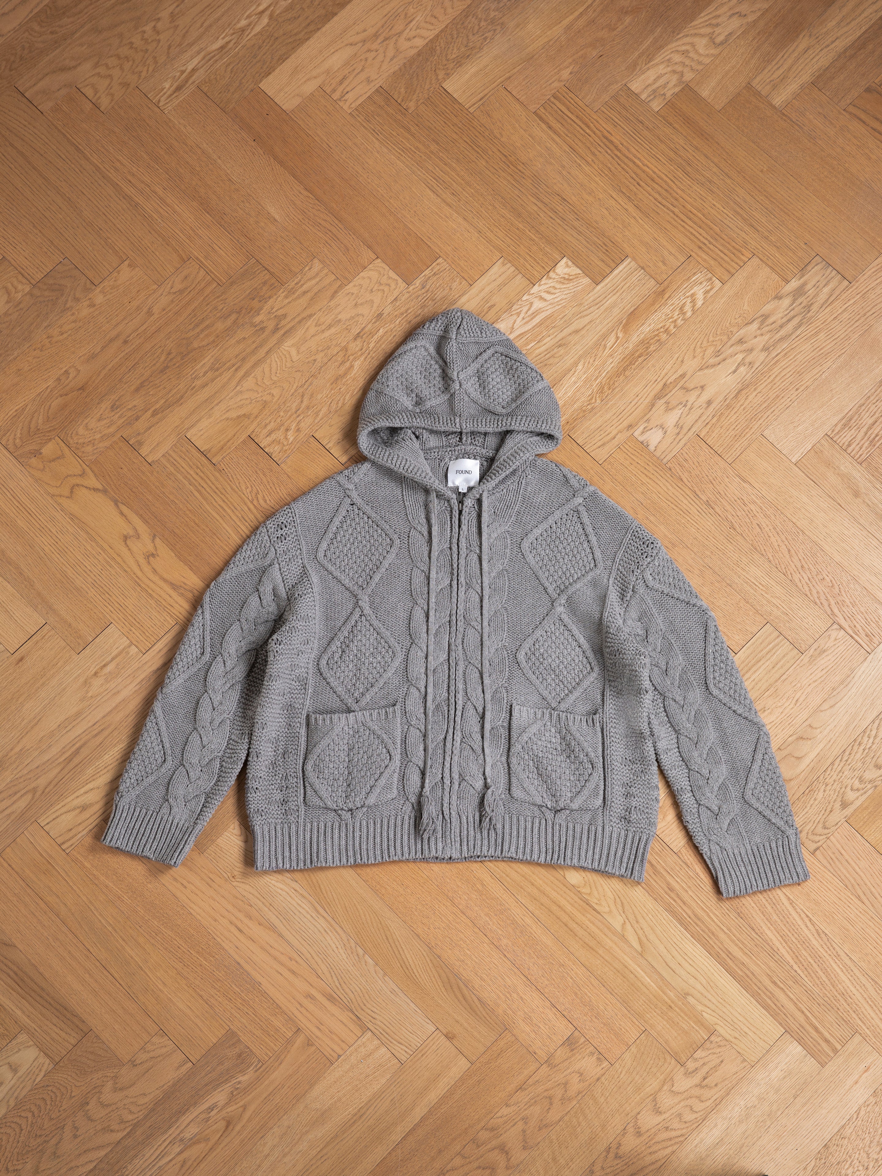 A gray Found Zip Up Cable Knit Hoodie is laid flat on a herringbone wooden floor, showcasing its chunky knit design that promises comfort and warmth.